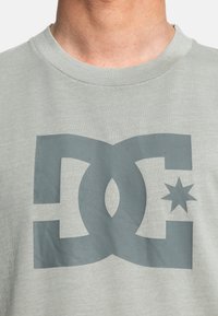Světle šedé bavlněné tričko s velkým tmavě šedým logem DC, doplněné designem hvězdy. Klasický crew výstřih s proužkovanou texturou.