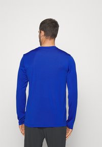 Man i en enkel blå långärmad skjorta och mörka byxor, vänd bort mot en enkel ljusgrå bakgrund.