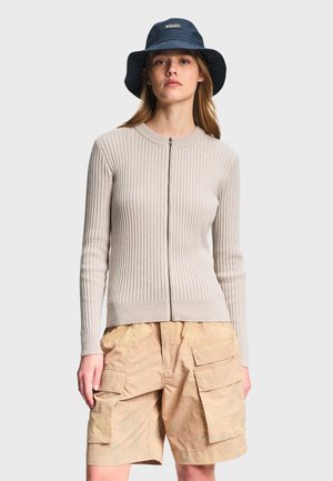 Ung kvinde iført navy bøllehat, beige ribstrikket cardigan med lynlås og brune cargoshorts, stående foran en lys baggrund.