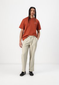 Tricou cu mânecă scurtă de culoare ruginită, cu un grafic subtil, asociat cu pantaloni plisati de culoare bej deschis și adidași negri.