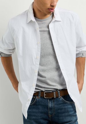 Man draagt een open witte overhemd over een grijs t-shirt, blauwe spijkerbroek en een bruine riem, met handen in de zakken tegen een witte achtergrond.