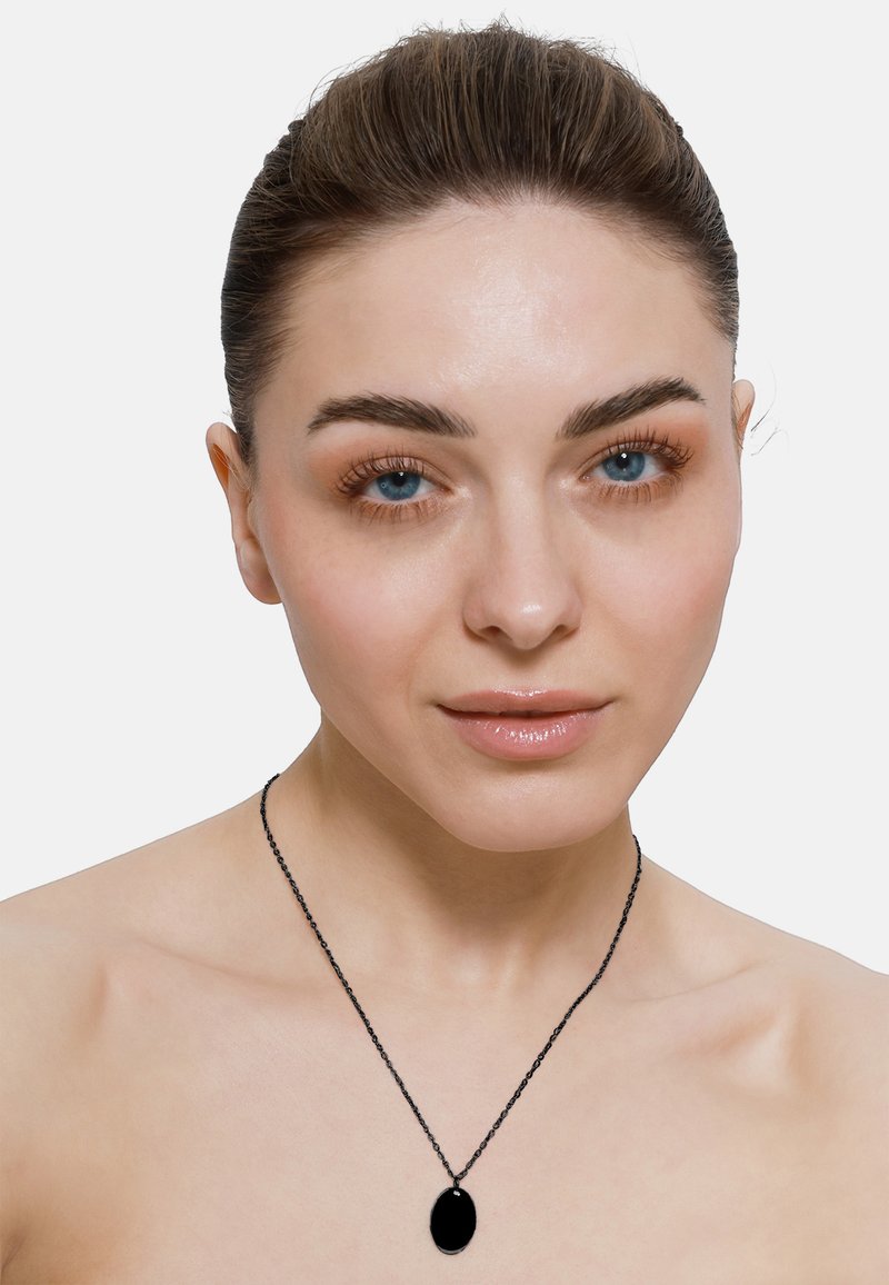 Collier avec pendentif ovale noir sur une chaîne fine, reposant sur une peau nue. Le pendentif présente une surface lisse et polie avec une finition brillante.