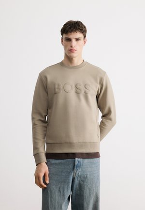 Mladý muž v béžovej mikine s nápisom "BOSS" na prednej strane a svetlomodrých džínsoch stojí pred jednoduchým bielym pozadím.