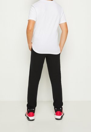 Niño de pie de espaldas, vistiendo una camiseta blanca lisa, pantalones negros y zapatillas rojas, negras y blancas, con las manos en los bolsillos.