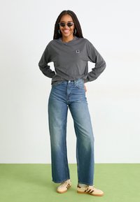 Chemise grise à manches longues avec un petit logo, associée à un jean bleu à jambes larges et des baskets beiges avec des accents bruns, se tenant sur un sol vert.