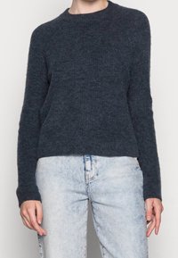 Pull en maille bleu marine foncé avec un col rond, des manches longues et une texture côtelée. Associé à un jean bleu clair déchiré.