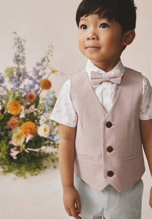 Ung dreng iført en lys lyserød vest, butterfly og blomstrede skjorte står ved siden af en farverig blomsterdekoration på en lys baggrund.