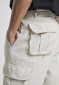 Beige cargobyxor med en mjuk, texturerad tygkonstruktion, stora sidofickor och en avslappnad passform. Detaljer inkluderar synlig sömnad och en looper i midjan.