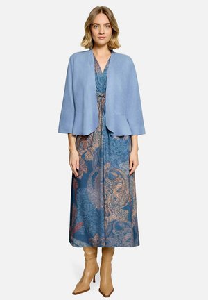 Vrouw met een blauwe paisley jurk, lichtblauwe open jas met driekwart mouwen en beige hoge hakken laarzen staand tegen een witte achtergrond.