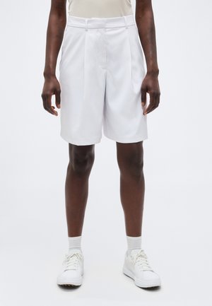 Osoba ubrana w białe, dopasowane szorty z wysokim stanem, białe skarpetki i białe trampki, stojąca na tle jednolitego jasnego tła.