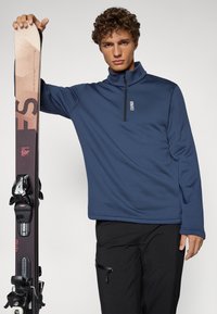 Top in pile blu navy con zip a metà altezza e colletto alto, abbinato a pantaloni da sci neri e a uno sci con grafiche multicolori e attacchi laterali.
