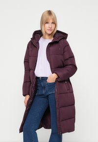 Dlhý, páperový kabát v tmavofialovej farbe s kapucňou. Obsahuje zips a bočné vrecká, skombinovaný s ľahkým topom a modrými džínsami. Hladká textúra.