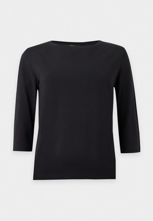 Femme debout portant un haut noir à manches trois-quarts, un jean gris foncé et des ballerines Mary Jane rouge foncé brillantes sur un fond uni.
