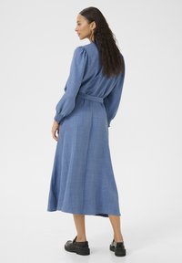 Robe longue en denim bleu à manches longues avec un détail smocké au niveau des manches et une ceinture à la taille, présentant une jupe fluide et des chaussures noires plates.