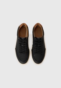Black sneakers made of leather with a textured finish, brown accents on the heel, flat laces, and perforated toe design.