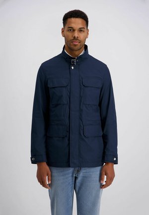 Mann trägt eine marineblaue Jacke mit vier vorderen Klappentaschen und einem Kragen mit Schnalle, kombiniert mit hellblauen Jeans, vor hellem Hintergrund.