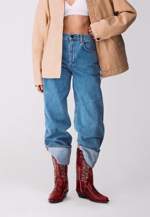 Persoon die een blauw, hooggetailleerd spijkerbroek draagt die in rode, geborduurde cowboylaarzen is gestopt, een wit crop top en een beige jas.