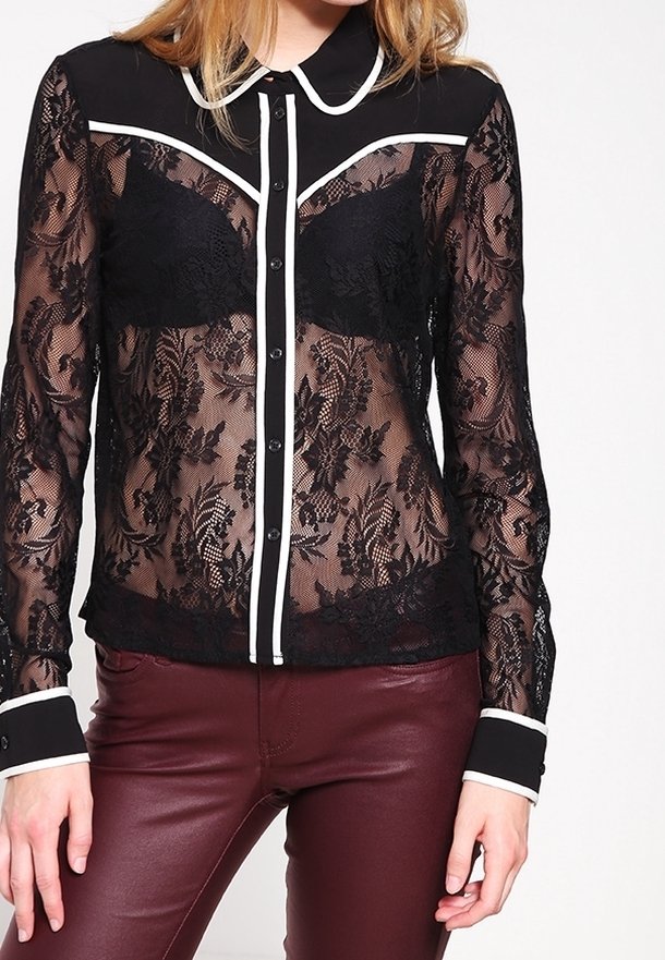 Femme portant un chemisier en dentelle noire transparente à boutons avec bordure blanche et un pantalon en cuir bordeaux sur un fond uni.