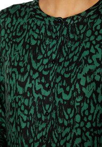 Tela con un patrón verde y negro con diseño de estampado de leopardo. Presenta un escote redondeado y botones negros en la parte delantera.