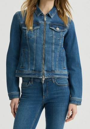 Femme portant une veste en jean bleue zippée avec des poches poitrine boutonnées et un jean bleu assorti, debout, les mains détendues.