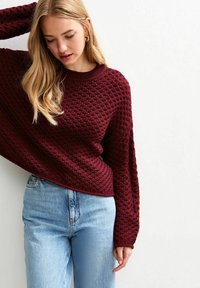 Vrouw met lang blond haar, gekleed in gouden ringoorbellen, een gestructureerde donkerrode trui en lichtblauwe jeans, staand tegen een witte achtergrond.
