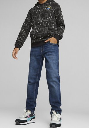 Personne portant un sweat à capuche noir Puma avec des logos, un jean bleu et des baskets blanches avec des accents noirs et turquoise, debout devant un fond gris.