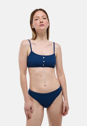 Femme à la peau claire et aux cheveux lisses châtain clair portant un bikini côtelé bleu marine avec trois boutons sur le devant du haut.