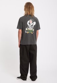 Personne aux cheveux longs, de dos, portant un t-shirt sombre avec un motif coloré de squelette tenant un bol vert dans le dos.