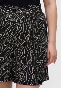 Short noir avec des motifs de lignes ondulées blanches porté par une personne avec un haut noir, montrant la taille et le bras droit.
