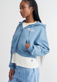 Corta azul em capuz feito de tecido macio, com bolsos frontais, capuz com cordão e texto bordado. Usado sobre uma camisa branca.