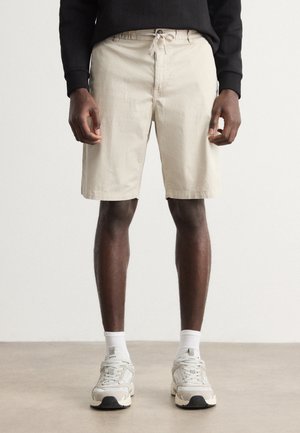 Short en coton beige avec une ceinture élastique et un cordon de serrage, présentant une coupe droite et des poches standard, accompagné de baskets grises.