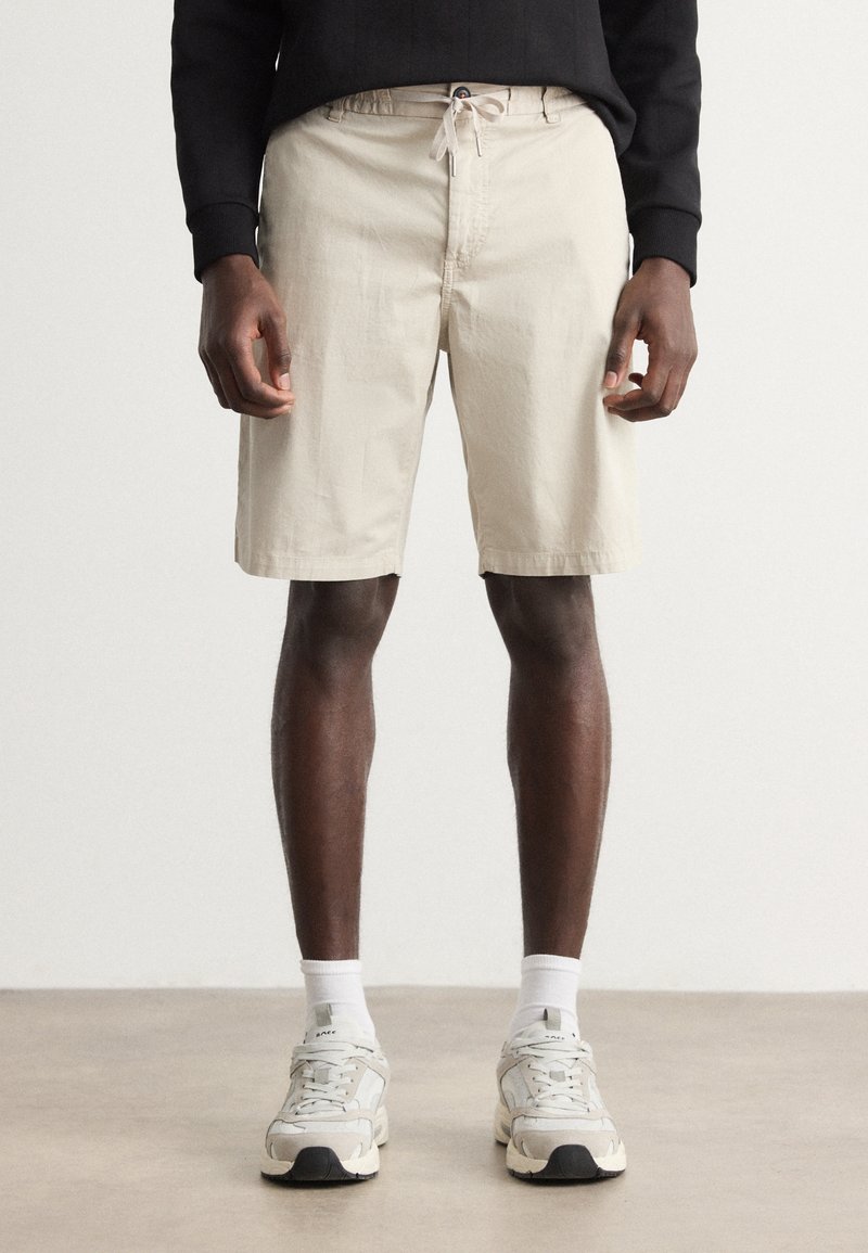 Short en coton beige avec une ceinture élastique et un cordon de serrage, présentant une coupe droite et des poches standard, accompagné de baskets grises.