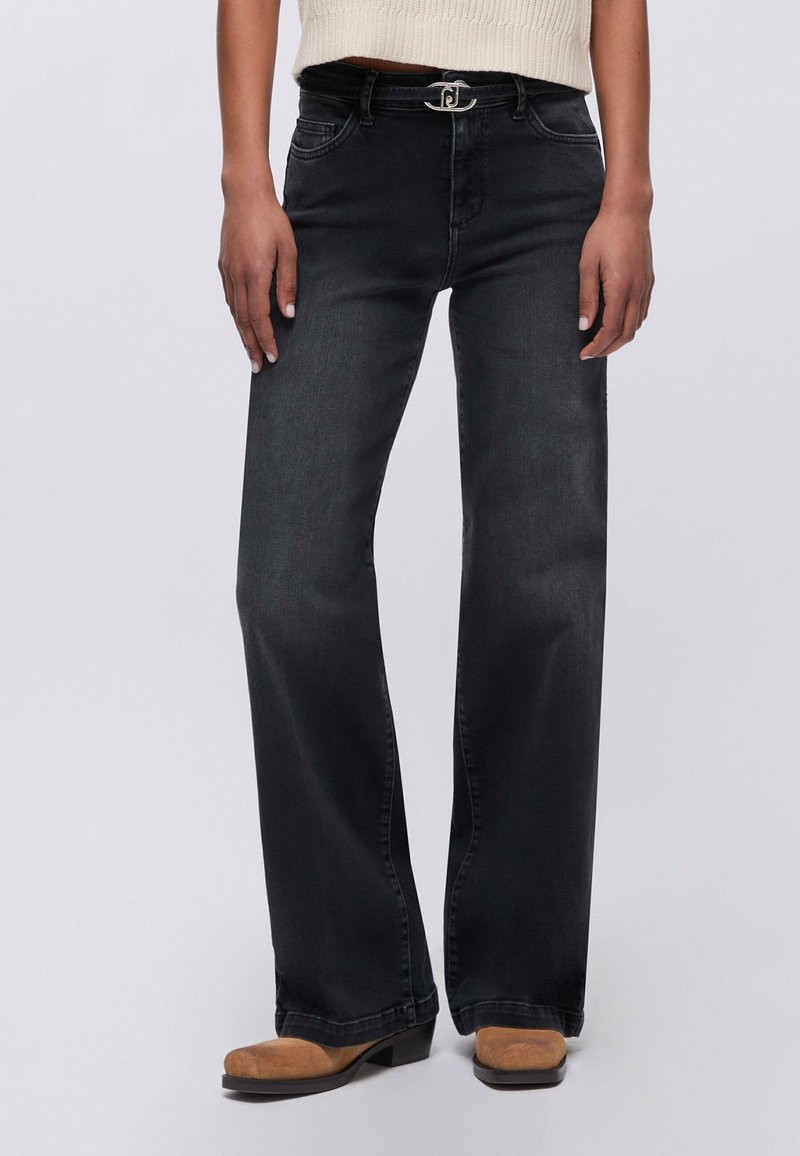 Femme portant un jean noir à jambes larges avec un bouton argenté et des bottes marron à bout carré, debout devant un fond clair.