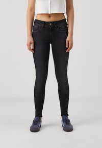 Vaqueros negros ajustados con una textura de denim suave, que presentan un diseño clásico de cinco bolsillos y un cierre de botón. Llevados con zapatillas deportivas azules.