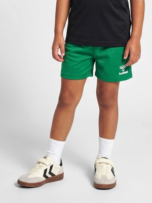 Barn iført grønne sportsshorts, hvide sokker og cremefarvede sneakers med sorte chevron-detaljer på en ensfarvet baggrund.