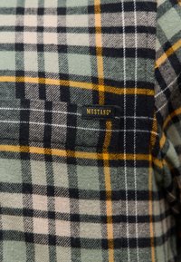 Grünes kariertes Flanellhemd mit gelben und schwarzen Akzenten. Verfügt über ein angenähtes Etikett mit der Aufschrift "MUSTANG®" in Schwarz auf dunklem Hintergrund.