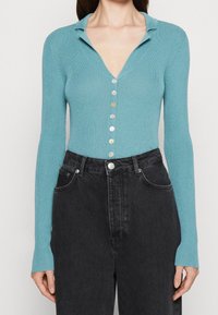 Cardigan rigato turchese con scollatura a V profonda, maniche lunghe e sette bottoni di colori vari, abbinato a jeans neri a vita alta.