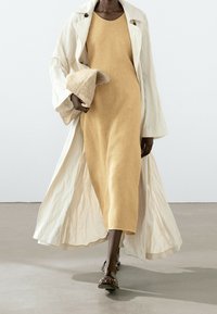Langer, crema-farbener Mantel über einem senfgelben Leinenkleid. Eine strukturierte Strohtasche wird in einer Hand gehalten. Flache Sandalen vervollständigen das Outfit.