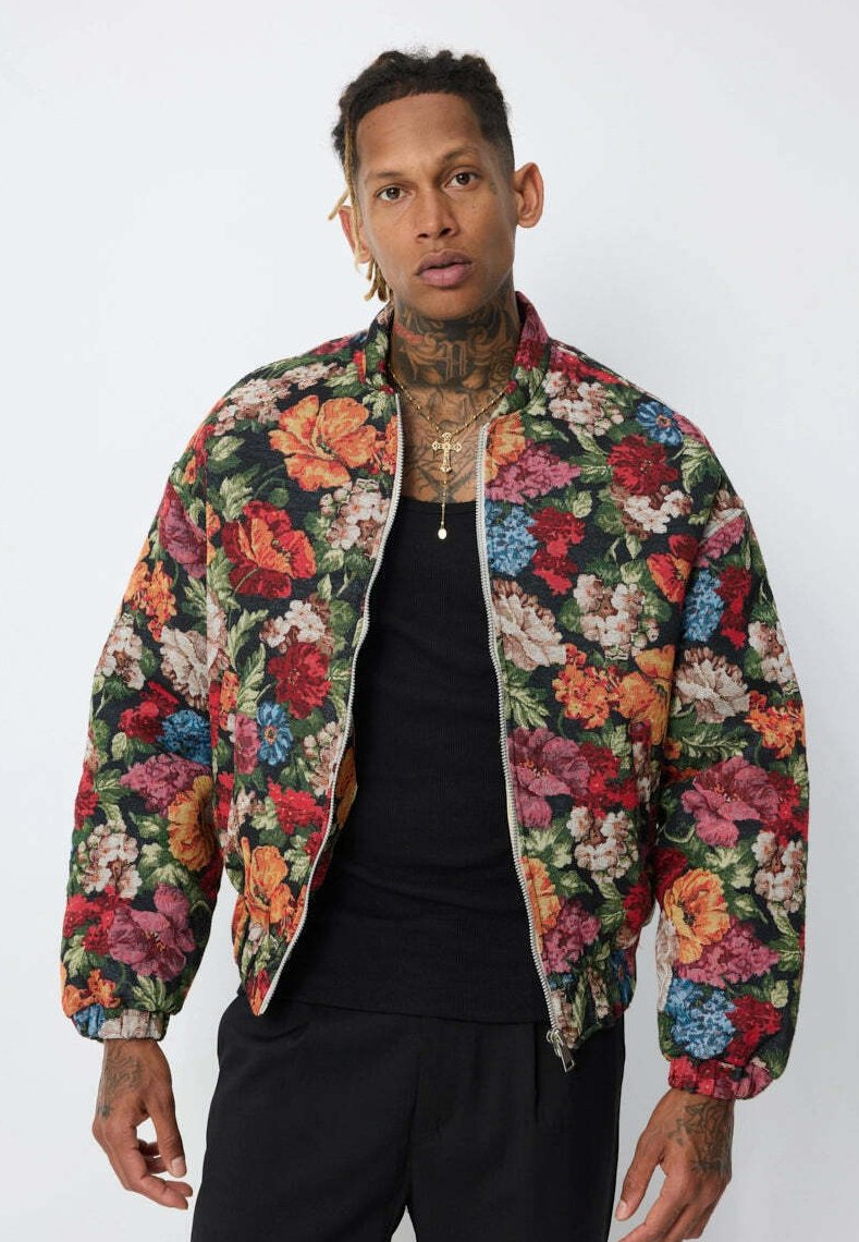 Homme avec des tatouages au cou et aux mains portant une veste zippée à motifs floraux colorés par-dessus un t-shirt noir, se tenant devant un fond uni.