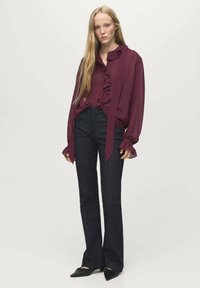 Blouse bordeaux avec détail volanté et manches longues ; associé à un jean évasé noir et des chaussures noires à bout pointu. Design propre et simple.