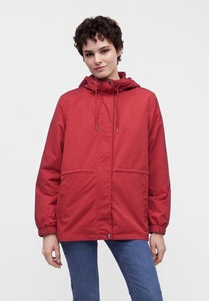 Jeune personne avec des cheveux courts et bouclés portant une veste à capuche rouge et un jean bleu sur un fond blanc uni.