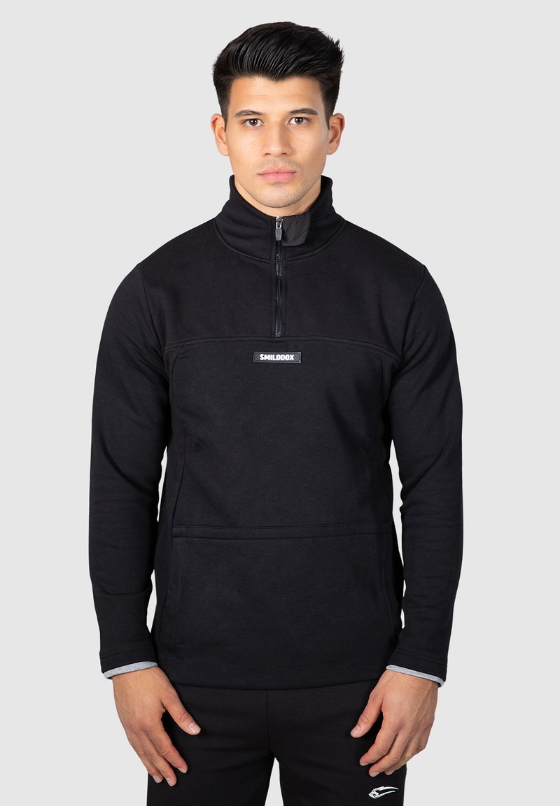 Mustavillainen fleece-pullove, jossa on korkea kaulus, vetoketju ja etutasku. Rinnassa on logopatch ja hihansuissa on ribbivarsia.