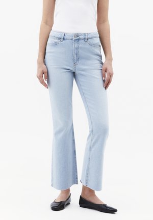 Frau trägt hellblaue, kurze Schlagjeans, ein weißes ärmelloses Oberteil und schwarze flache Schuhe, steht vor weißem Hintergrund.