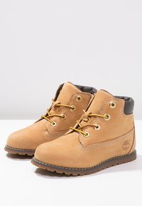 Botas de ante marrón con ojales dorados y cordones amarillos. Presentan un cuello acolchado y una suela de goma. Logotipo grabado en el lateral.