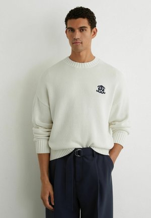 Hombre con suéter de punto blanco con logo bordado en negro y pantalones azul marino con cinturón de tela, de pie frente a una pared lisa y clara.