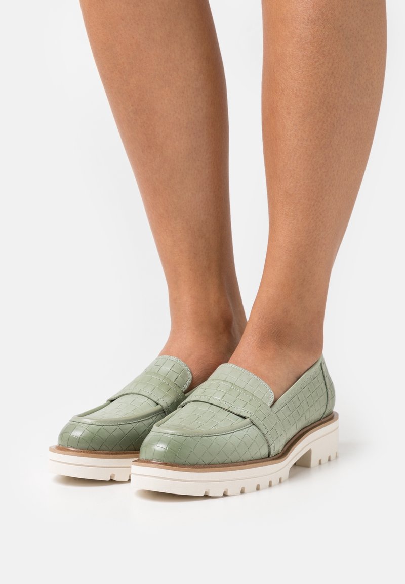 Pieds portant des mocassins en cuir tressé vert clair avec des semelles blanches épaisses sur un fond clair.