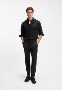 Μαύρο πουκάμισο με κουμπιά και κεντημένο λογότυπο, συνδυασμένο με στενά μαύρα παντελόνια και μαύρα αθλητικά παπούτσια. Το μοντέλο στέκεται με τα χέρια στις τσέπες.