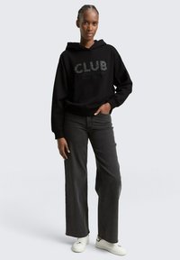 Schwarzer Kapuzenpullover mit "CLUB"-Text und Logo, kombiniert mit dunkelgrauen weiten Jeans und weißen Turnschuhen. Glatter Stoff und lässige Passform.