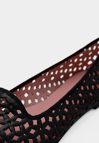 Μαύρο επίπεδο παπούτσι με περίπλοκο γεωμετρικό μοτίβο κοπής και απαλό ροζ εσωτερικό πέλμα, σε λοξή όψη σε λευκό φόντο.