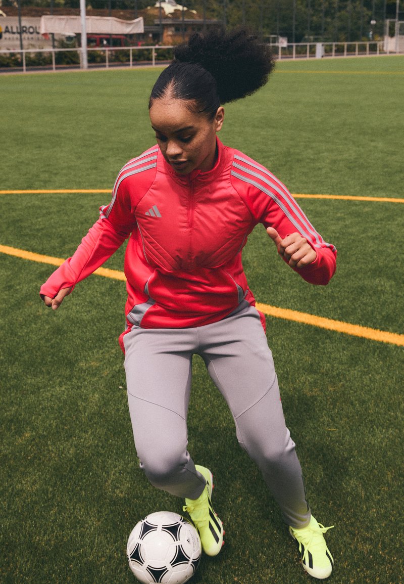 Rote Sportjacke mit grauen Akzenten, graue Hose und leuchtend gelbe Fußballschuhe. Fußball auf grünem Rasen in der Nähe der Füße der Person.